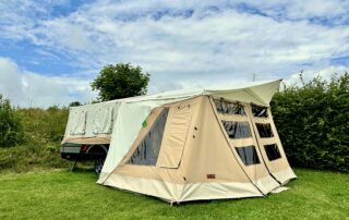 Faltcaravan Combi-Camp Country mit Vorzelt in Beige-Ecru auf einer grünen Wiese mit blauem wolkendurchzogenem Himmel. Eine Hecke und ein Baum im Hintergrund